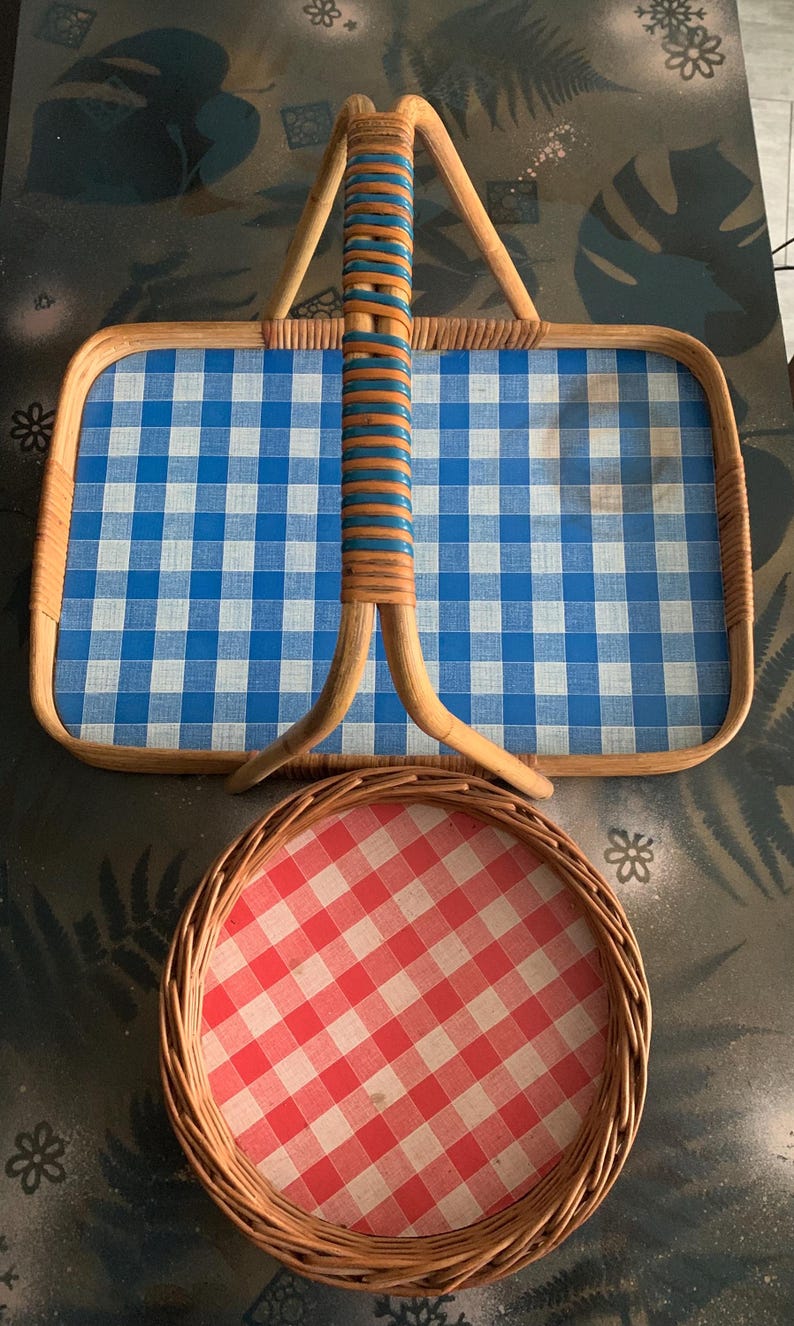 May include: Two picnic baskets. The larger basket is rectangular with a blue and white checkered pattern and a woven handle with blue and brown accents. The smaller basket is round with a red and white checkered pattern.