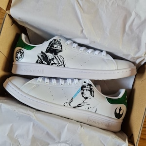 Pode incluir: Um par de t&ecirc;nis brancos Adidas Stan Smith com detalhes verdes. O t&ecirc;nis esquerdo apresenta uma ilustra&ccedil;&atilde;o em preto e branco de Darth Vader de Star Wars, enquanto o t&ecirc;nis direito apresenta uma ilustra&ccedil;&atilde;o em preto e branco de Luke Skywalker de Star Wars. Os t&ecirc;nis est&atilde;o em uma caixa com papel de seda branco.