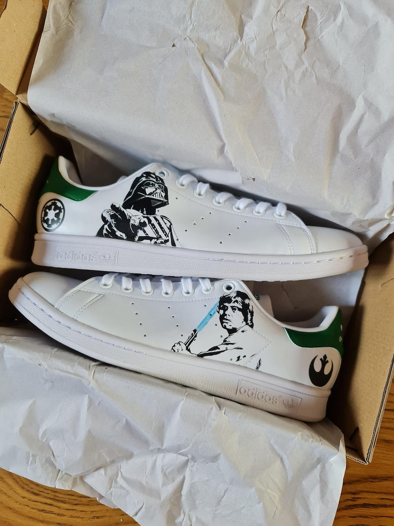 Pode incluir: Um par de t&ecirc;nis brancos Adidas Stan Smith com detalhes verdes. O t&ecirc;nis esquerdo apresenta uma ilustra&ccedil;&atilde;o em preto e branco de Darth Vader de Star Wars, enquanto o t&ecirc;nis direito apresenta uma ilustra&ccedil;&atilde;o em preto e branco de Luke Skywalker de Star Wars. Os t&ecirc;nis est&atilde;o em uma caixa com papel de seda branco.