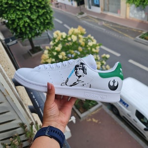 Pode incluir: T&ecirc;nis Adidas Stan Smith branco com acabamento verde e um design personalizado que apresenta uma imagem em preto e branco de Luke Skywalker de Star Wars segurando um sabre de luz azul. O s&iacute;mbolo da Alian&ccedil;a Rebelde est&aacute; no calcanhar.