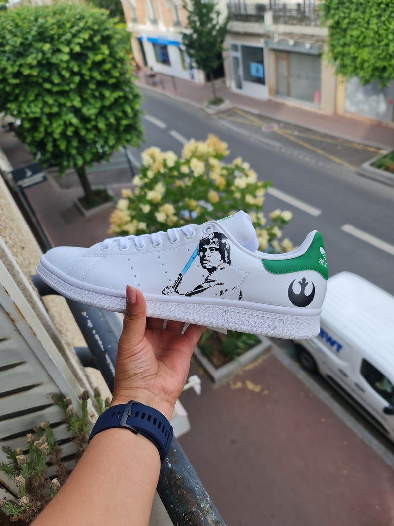 Pode incluir: T&ecirc;nis Adidas Stan Smith branco com acabamento verde e um design personalizado que apresenta uma imagem em preto e branco de Luke Skywalker de Star Wars segurando um sabre de luz azul. O s&iacute;mbolo da Alian&ccedil;a Rebelde est&aacute; no calcanhar.