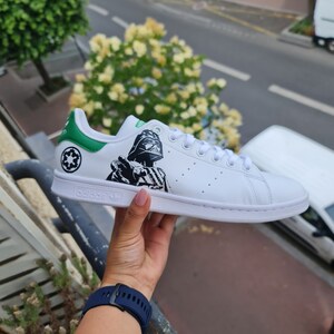 Pode incluir: T&ecirc;nis branco Adidas Stan Smith com acabamento verde e um gr&aacute;fico preto e branco do Darth Vader no lado. O texto "adidas" &eacute; impresso no lado do t&ecirc;nis.