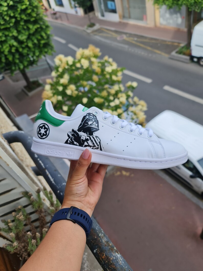 Pode incluir: T&ecirc;nis branco Adidas Stan Smith com acabamento verde e um gr&aacute;fico preto e branco do Darth Vader no lado. O texto "adidas" &eacute; impresso no lado do t&ecirc;nis.