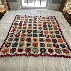 May include: A colourful, handmade crochet blanket with a white base and floral-style squares in shades of orange, red, green, and blue. The blanket has a dark red border with small pom-poms around the edge.