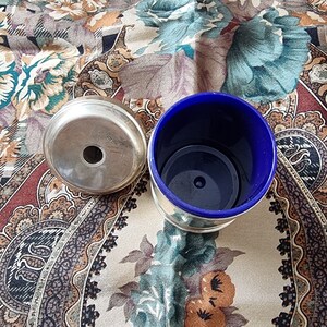 K&ouml;nnte beinhalten: Ein silberfarbener Aschenbecher mit einem zentralen Loch liegt neben einem blau-silbernen Beh&auml;lter. Der Beh&auml;lter hat eine blaue Innenseite und einen silbernen Rand. Die Artikel befinden sich auf einem gemusterten Stoff mit Blumen- und Paisley-Designs.