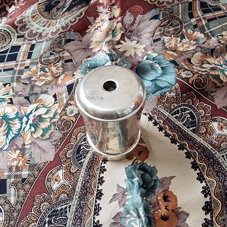 K&ouml;nnte beinhalten: Ein silberfarbener, zylindrischer Beh&auml;lter mit einem Loch in der Mitte des Deckels. Der Beh&auml;lter ist reflektierend und liegt auf einem gemusterten Stoff mit Blumen- und Paisley-Designs in Braun-, Blau- und Cremet&ouml;nen.