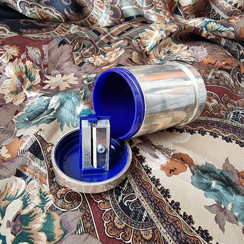 K&ouml;nnte beinhalten: Ein silberfarbener Bleistiftanspitzer mit blauem Innenraum und passendem blauem Deckel. Der Anspitzer ist zylindrisch und hat ein silbernes Band in der Mitte. Der Deckel ist ge&ouml;ffnet und zeigt den Anspitzer und seine Komponenten.