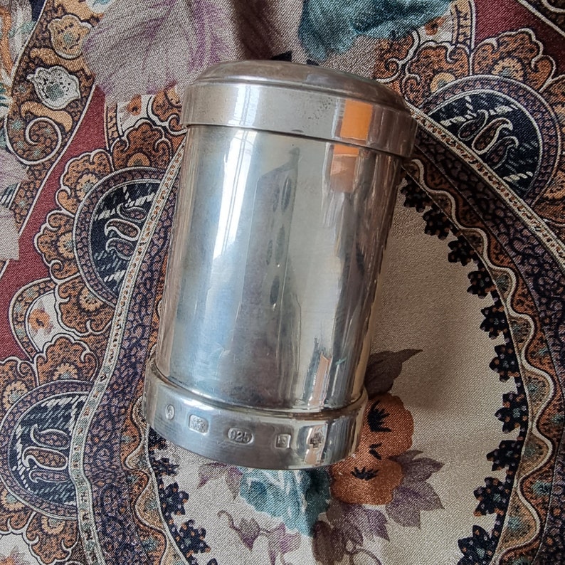 K&ouml;nnte beinhalten: Ein zylindrischer Silberbeh&auml;lter mit Deckel. Der Beh&auml;lter besteht aus einem gl&auml;nzenden, reflektierenden Metall. Der Boden des Beh&auml;lters hat mehrere kleine Markierungen. Der Beh&auml;lter steht auf einem Stoff mit Blumen- und Paisley-Mustern.