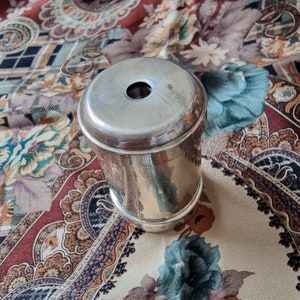 K&ouml;nnte beinhalten: Ein silberfarbener, zylindrischer Beh&auml;lter mit Deckel. Der Beh&auml;lter hat ein kleines Loch in der Mitte des Deckels. Die Oberfl&auml;che des Beh&auml;lters reflektiert die Umgebung. Der Beh&auml;lter steht auf einem gemusterten Stoff.