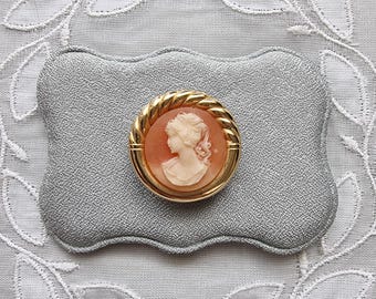 May include: A cameo brooch with a gold rope-like frame and a white cameo of a woman's profile. The cameo is set against a peach-colored background.