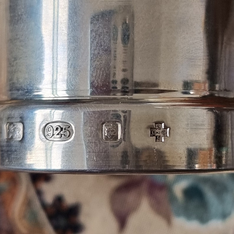 K&ouml;nnte beinhalten: Nahaufnahme eines polierten Silberobjekts mit mehreren Punzierungen. Die Punzierungen umfassen einen Stempel "925", der Sterlingsilber anzeigt, und andere komplizierte Symbole. Die Oberfl&auml;che reflektiert das Licht und hebt die glatte Textur und Handwerkskunst des St&uuml;cks hervor.