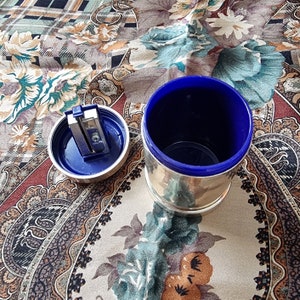 K&ouml;nnte beinhalten: Ein silberfarbener Bleistiftanspitzer mit blauem Innenraum und passendem Deckel. Der Deckel ist ge&ouml;ffnet und zeigt die Anspitzerklinge. Der zylindrische Beh&auml;lter steht auf einem gemusterten Stoff.