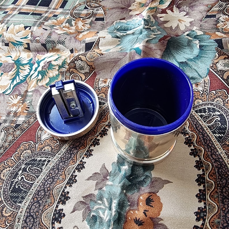 K&ouml;nnte beinhalten: Ein silberfarbener Bleistiftanspitzer mit blauem Innenraum und passendem Deckel. Der Deckel ist ge&ouml;ffnet und zeigt die Anspitzerklinge. Der zylindrische Beh&auml;lter steht auf einem gemusterten Stoff.