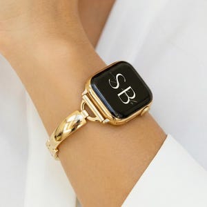 Puede incluir: Un Apple Watch dorado con una pantalla negra que muestra las iniciales "SB" en blanco. El reloj está sujeto a una correa dorada a juego con un diseño tejido. El reloj se lleva en la muñeca, sobre un fondo blanco.
