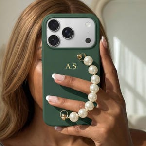 May include: A dark green phone case with a pearl wrist strap. The case has the initials "A.S" in gold lettering. The phone case is being held up to a mirror.