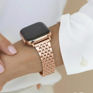 Puede incluir: Reloj de oro rosa con esfera rectangular y correa de eslabones a juego. El reloj se lleva en la muñeca, con una manga de camisa blanca visible en el fondo. La correa del reloj mide aproximadamente 18 cm de largo.