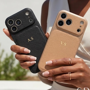 May include: Two personalized phone cases, one black and one tan, are held up. The black case has the initials "A.S" in gold, while the tan case has "V.S". Both cases have a textured leather appearance and a camera module.
