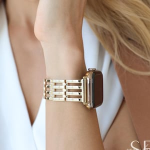 Puede incluir: Correa de reloj dorada con un diseño rectangular. La correa está sujeta a un reloj dorado con pantalla oscura. La correa del reloj tiene una serie de barras horizontales. El reloj está en una muñeca.