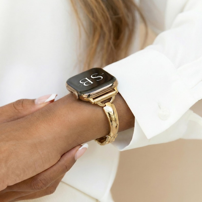 May include: Gold-toned smartwatch with a matching band. The watch face displays the initials "SB" in white. The band has a textured design and is secured on the wrist. The watch is worn with a white long-sleeved shirt.