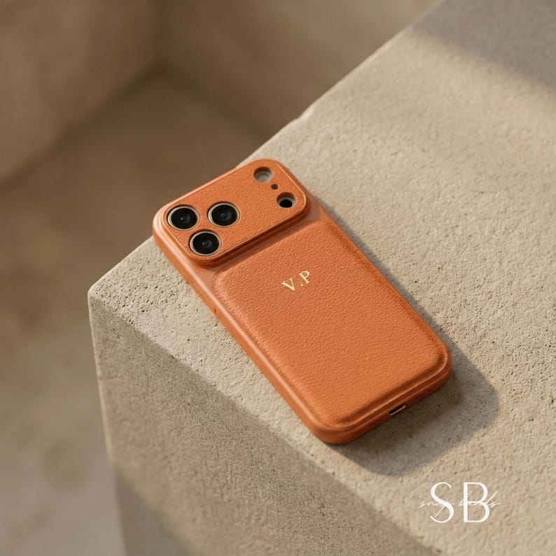 May include: A tan leather phone case with the initials "V.P" embossed on the back. The case has a camera cutout and is designed to fit a smartphone. The case is sitting on a light-colored surface.