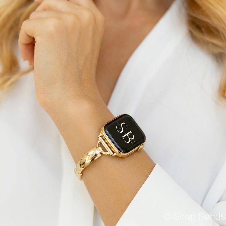 May include: A gold-toned Apple Watch with a black screen displaying the initials "SB" in white. The watch is attached to a matching gold band with a woven design. The watch is worn on a wrist, against a white background.