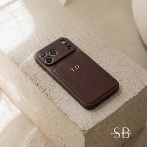 May include: A brown leather phone case with a camera cutout and the initials "T.D" in gold lettering. The case is on a textured, light-colored surface. The case has a smooth, leather-like texture.