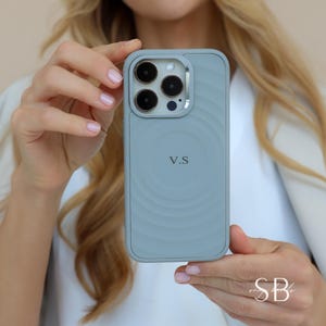 Puede incluir: Una funda de tel&eacute;fono azul claro con un dise&ntilde;o circular en la parte posterior. La funda tiene un recorte para la c&aacute;mara y las iniciales "V.S" est&aacute;n impresas en la parte posterior. La funda est&aacute; sujeta por una persona.