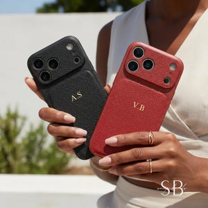 May include: Two phone cases, one black and one red, are held up. The black case has the initials "A.S" in gold, and the red case has "V.B". Both cases have a textured leather appearance and are designed for a smartphone.