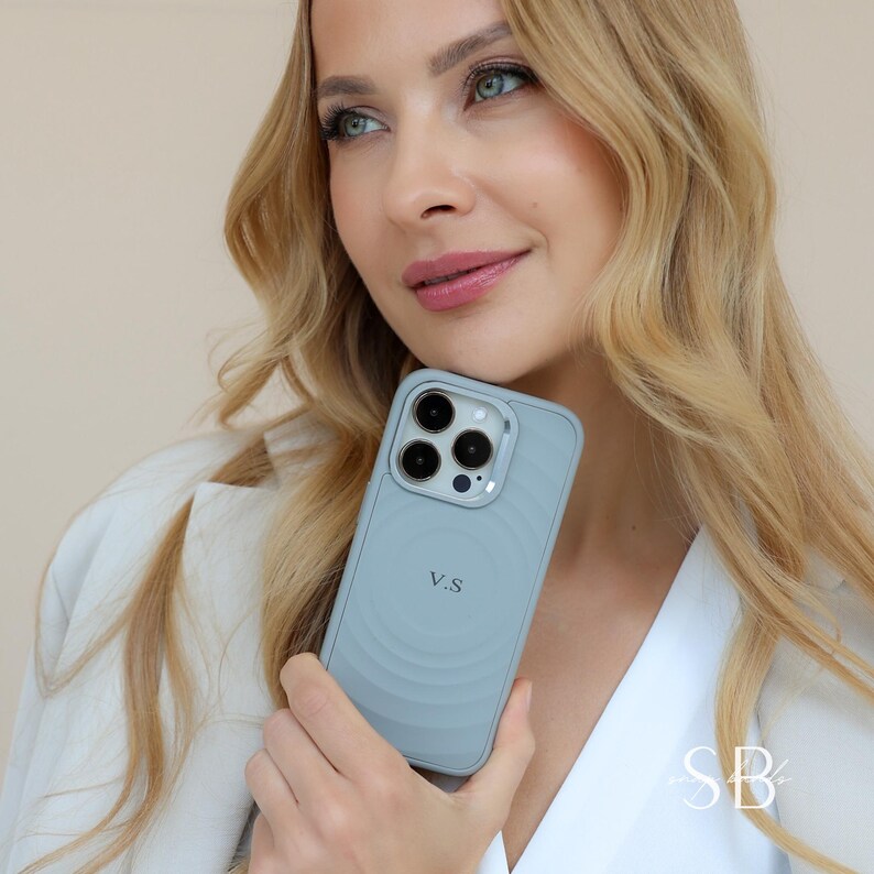 Puede incluir: Una funda de tel&eacute;fono azul claro con un dise&ntilde;o circular y las iniciales "V.S" se muestra. La funda tiene un parachoques protector y un recorte para la c&aacute;mara. La persona lleva una blazer blanca.