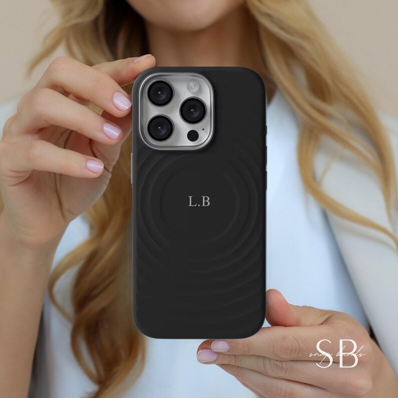 Puede incluir: Funda de tel&eacute;fono negra con un dise&ntilde;o circular y las iniciales "L.B" en el centro. La funda tiene un anillo de c&aacute;mara plateado. La persona que sostiene el tel&eacute;fono tiene el pelo rubio.