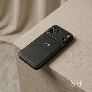 May include: Black leather phone case with gold lettering "A.S". The case has a textured surface and a camera cutout. The phone case is on a neutral-colored surface.