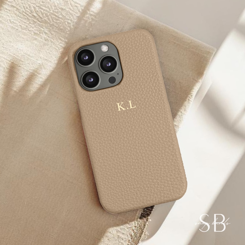 May include: A beige leather phone case with a gold monogram "K.L." on the back. The case is designed for an iPhone with a triple lens camera.