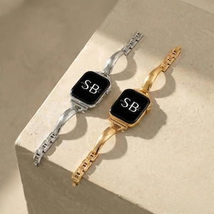 May include: Two smartwatches with black screens, one silver and one gold. Each watch displays the initials "SB" in white. The bands are made of linked metal segments and curved, solid metal. The watches are angled on a light-colored surface.