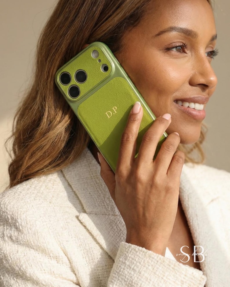 May include: A green phone case with the initials "D.P." in gold lettering. The case is held up to the ear. The phone case has a textured leather-like appearance and a camera cutout.