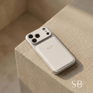 May include: A light gray phone case with a leather texture. The case has a camera cutout and the initials "G.P" in gold lettering. The case is resting on a light-colored surface.