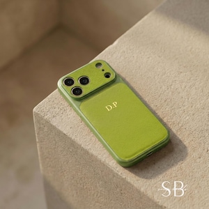 May include: A green phone case with a textured leather-like finish. The case has a camera cutout and the initials "D.P" in gold. The case is on a neutral-toned surface.