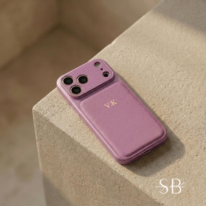 May include: A mauve-colored phone case with a textured finish. The case has a camera cutout and the initials "V.K." in gold lettering. The case is resting on a light-colored surface.