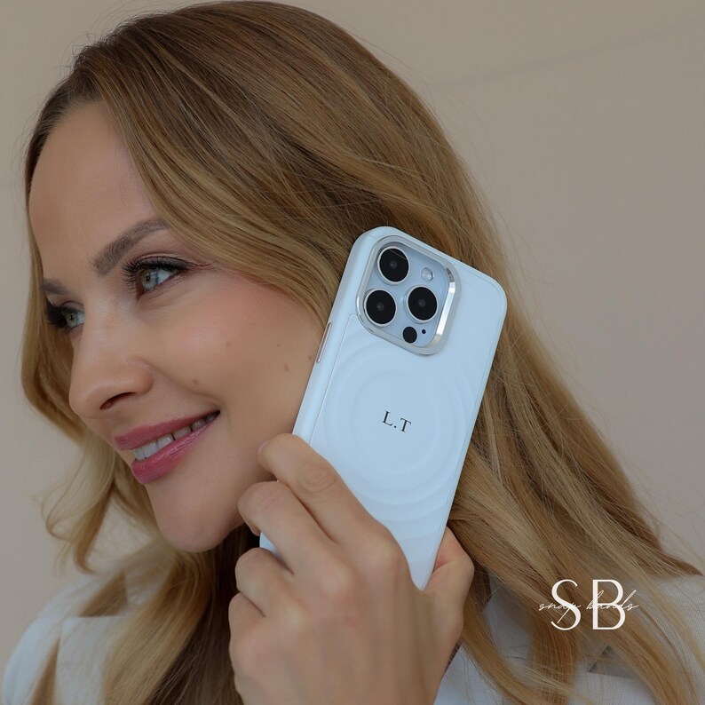 Puede incluir: Una funda de tel&eacute;fono blanca con un dise&ntilde;o circular y las iniciales "L.T" impresas en la parte posterior. La funda tiene un anillo de c&aacute;mara plateado. La funda se sostiene frente a una persona con cabello rubio.