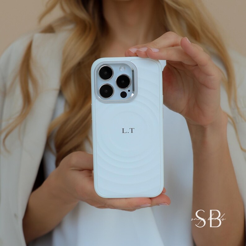 Puede incluir: Funda de tel&eacute;fono azul claro con un anillo de c&aacute;mara plateado y las iniciales "L.T" impresas en la parte delantera. La funda tiene un dise&ntilde;o texturizado circular. La funda del tel&eacute;fono se sostiene frente a una persona.