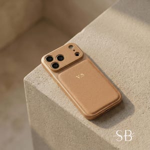 May include: A tan leather phone case with the initials "V.S." in gold lettering. The case has a textured surface and a camera cutout. The case is on a light-colored surface. The letters "SB" are in the bottom right corner.
