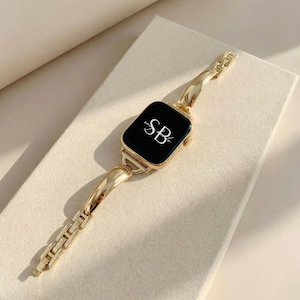 May include: Gold-toned smartwatch with a black screen displaying the initials "SB" in white script. The watch features a matching gold band with a combination of curved and rectangular links. The watch is set against a neutral background.