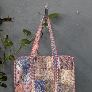 May include: A patchwork tote bag with a zippered pocket. The bag is made of cotton fabric and has a pink and white striped trim. The bag is hanging on a hook.