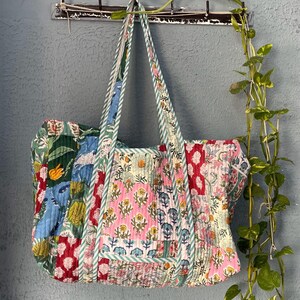 May include: A patchwork tote bag with floral and geometric patterns in shades of pink, green, blue, and red. The bag has two long straps and is hanging on a hook.