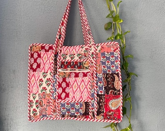 May include: A patchwork tote bag with a red, white, and blue trim. The bag is made of different fabrics with floral and geometric patterns. It is hanging on a hook with a vine plant in the background.