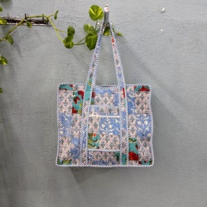 Op de afbeelding: Een blauwe en witte patchwork tote bag met een bloemenprint en een kleiner vakje aan de voorkant. De tas hangt aan een haak aan een grijze muur.