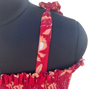 May include: Red sundress with floral print and tie straps. The dress features a smocked bodice and a vibrant pattern of white, orange, and maroon flowers. The straps are tied at the shoulders, adding a delicate touch to the summer dress.