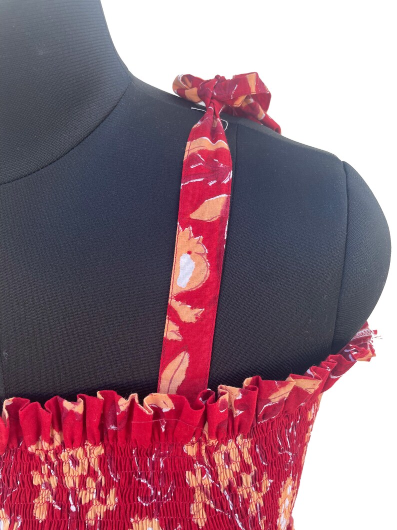 May include: Red sundress with floral print and tie straps. The dress features a smocked bodice and a vibrant pattern of white, orange, and maroon flowers. The straps are tied at the shoulders, adding a delicate touch to the summer dress.