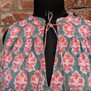 May include: A sleeveless green cotton top with a floral print in shades of pink and orange. The top has a V-neckline and a tie at the neck.