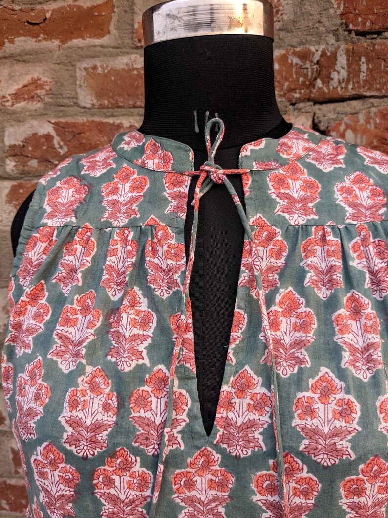 May include: A sleeveless green cotton top with a floral print in shades of pink and orange. The top has a V-neckline and a tie at the neck.