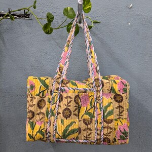 Könnte beinhalten: Eine gelbe und rosafarbene Baumwoll-Tragetasche mit floralem Muster und rosa-blauen, gestreiften Henkeln. Die Tasche hängt an einem Haken.